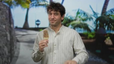 İspanyol adam dışarıda dondurma yerken, eğlenceli bir jest yapıyor, tropik bir sokak arkaplanına karşı kurulmuş, eğlence ve rahatlama karışımı yakalıyor..