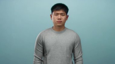 Gri kazak giyen genç adam, izole edilmiş mavi bir arka plana karşı duruyor, basit ve düşünceli bir ifadeyi yakalıyor..