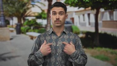 Genç İspanyol bir adam açık hava parkı ortamında sürpriz bir jest yapıyor, arka planda ağaçlar ve banklar olan desenli bir gömlek giyiyor, samimi bir kendini tanıma anını yakalıyor..