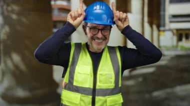 Kentsel sokaktaki İspanyol adam sıkı şapka ve güvenlik yeleği giyerek şakacı bir hareket yapıyor..