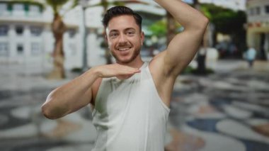 Sakallı genç adam kolsuz bir gömlek içinde gülümsüyor, enerjik bir şekilde hareketli bir şehir caddesinde arka planı bulanık.