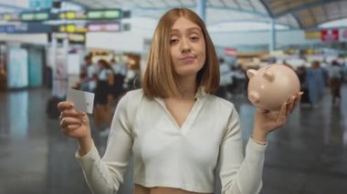 Havaalanı terminalinde elinde kumbara ve kredi kartı tutan bir kadın modern seyahat finansmanı ve dünya çapında genç gezginler için tasarruf stratejileri sergiliyor..
