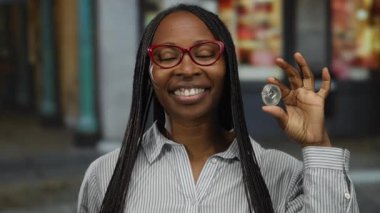 Şehir merkezinde elinde ethereum para tutan kadın şifreli para sergiliyor. Kendine güveni tam, örgülü saç stili, gözlük ve çizgili gömlek takıyor..