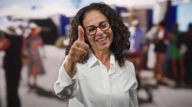 Kıvırcık saçlı bir kadın, kalabalık bir şehir caddesinde dikilirken, açık şehir ortamında kendine güveni ve mutluluğu yayıyor..