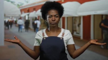 Önlüklü genç bir kadın, arka planda insanların olduğu düşünceli bir ifade sergiliyor..