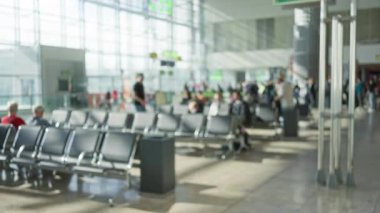 Havaalanı terminalinde bulanık bir sahne. Parlak ışık altında odakları kesilmiş insanlar ve boş sandalyeler..