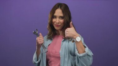 İngiliz anahtarı tutan İspanyol kadın, canlı mor bir arkaplan karşısında gülümseyerek, modern izole bir çevrede güven ve beceri ifade ediyor..