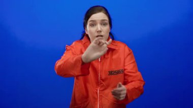 Mavi arka planda turuncu tulumlu kadın, kulakları ve jestleri kapatarak, kapalı bir ortamda duygusal yoğunluğu vurgulayarak hayal kırıklığını ifade ediyor..