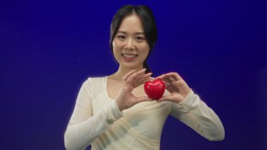 Kırmızı kalbi canlı mavi arka planda tutan bir kadın. Sıcak bir şekilde gülümsüyor. Aşkın ve pozitifliğin sıcaklığını dışa vuruyor.