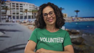 Orta yaşlı İspanyol gönüllü, deniz kenarındaki gezinti alanında kollarını kavuşturup gülümsüyor. Gönüllü hizmet nezaketi gösteriyor..