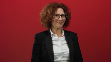 Orta yaşlı, takım elbiseli İspanyol bir kadın, canlı kırmızı bir duvara el hareketi yaparak izole edilmiş bir stüdyoda güven ve iletişimi harmanlıyor..