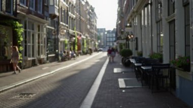 Amsterdam 'da güneşli bir kaldırım taşından yolda yürüyen ve şehir hayatını aydınlatan büyüleyici binalar ve kafelerle dolu bulanık bir sokak manzarası..