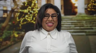 Gözlüklü genç İspanyol kadın şehir caddesinde rock jesti yapıyor. Arka planda neşeli ışıklar ve açık havada kendine güveni ifade ediyor..