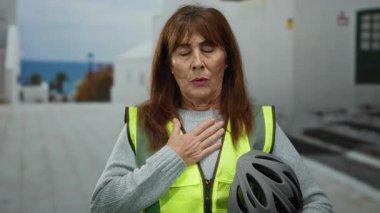 İspanyol kıdemli kadın şehir sokaklarında meditasyon yapıyor sarı bir yelek giyiyor, bisiklet kaskı tutuyor, elleri göğsünde, kentsel bir çevrede huzur ve sükunet gösteriyor..