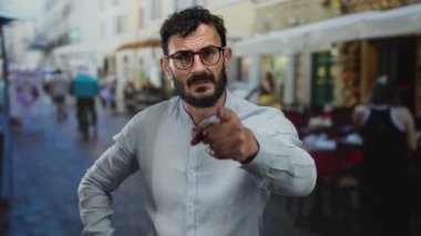 Canlı sokak sahnesinde sakallı İspanyol bir adam masaların ve yayaların hareketli kentsel geçmişleri arasında gözlük ve gri gömlek takıyor..