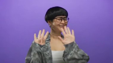 Neşeli bir şekilde el sallayan bir kadın canlı mor arka planda gözlük takıyor ve şık bir bluz giyiyor neşeli ve dostane bir tavır sergiliyor.