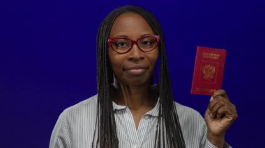 Mavi arka plan önünde Rus pasaportu taşıyan, gözlüklü ve çizgili gömlek giyen uluslararası seyahat ve kimlik kavramları taşıyan bir kadın..