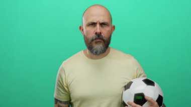 Futbol topu tutan sakallı kel bir adam, izole edilmiş yeşil arka plana karşı tehditkar hareketler yapıyor, duygularını ve yoğunluğunu açıkça ifade ediyor..