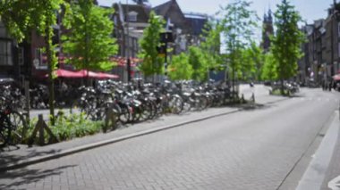Hollanda, Amsterdam 'da güneşli bir cadde boyunca dizilmiş bisikletler, ağaç astarlı yollar ve bulanık mimari yapılarla sakin bir kentsel atmosfer ve dağılmış bir ortam yaratıyor..