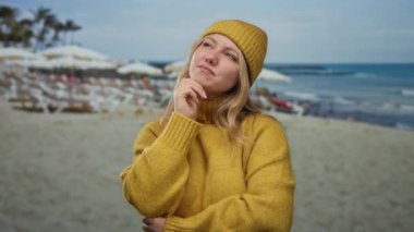 Sarı kazaklı ve şapkalı kadın bulutlu bir günde plajda bulanık bir deniz ve arka planda sandalyelerle dikiliyor..