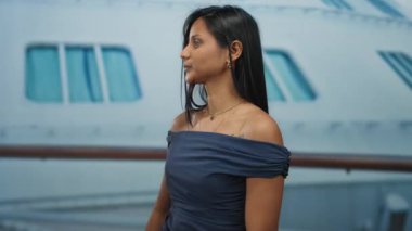 Yolcu gemisindeki genç bir kadın, denizcilik arkaplanına karşı mavi bir elbise ve altın takı takmış, el tabancasıyla neşeli poz veriyor..