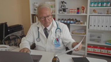 Beyaz üniformalı kıdemli erkek doktor hastane odasında tıbbi ekipman ve dizüstü bilgisayarla, odaklanmış ve profesyonelce evrak işlerini gözden geçiriyor..