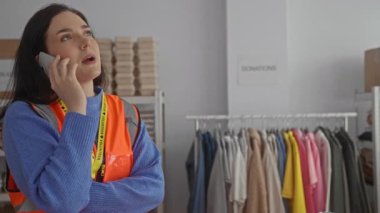 Bağış odasında, kıyafetlerle ve kutularla çevrili turuncu yelek giyen bir kadın telefonda konuşuyor..