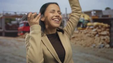 Bir inşaat alanında akıllı telefondan müzik dinlerken gülümseyen bir kadın, dışarıda çalışma ortamında neşe ve enerjiyi somutlaştırıyor..