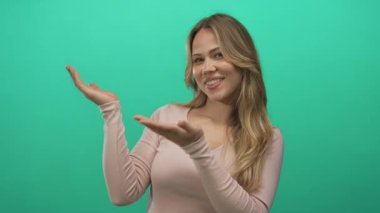 Sarışın, beyaz bir kadın yeşil bir stüdyoda gülümserken her iki avucunu havaya kaldırıyor. Uzun kollu bir bluzla; sıcak karşılama..