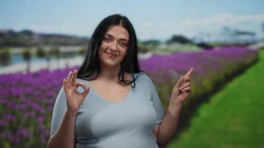 Canlı bir park ortamında kendine güvenen bir kadın, güzel bir jest yapıyor, kıvrımlı vücudunu vurguluyor, mor çiçekler ve açık havada neşeli bir hava için arka planda yeşillik var..