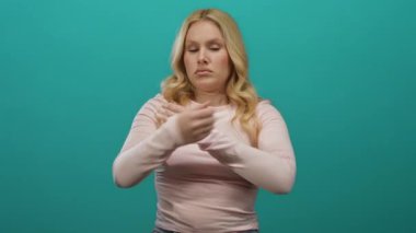 Sarı saçlı ve katlanmış kollu bir kadın pembe gömlek giyen canlı yeşil bir arka plana karşı güvenle duruyor..