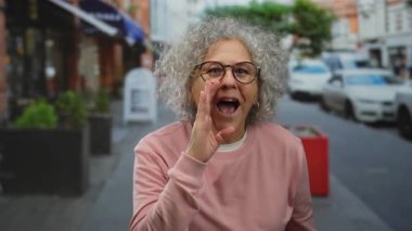 Şehir sokaklarında gülümseyen ve konuşan kır saçlı kadın, hareketli bir şehir ortamında olgun bir özgüvenin ve açık hava aktivitesinin özünü yakalıyor..