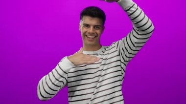 Çizgili tişörtlü, pembe arka planda elleri olan genç İspanyol adam neşeli ve neşeli bir ifade sergiliyor..