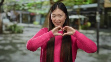 Pembe kazaklı bir kadın şehir sokaklarında elleri ile kalp şekli yapıyor. Neşenin, mutluluğun ve aşkın vücut bulmuş halidir..