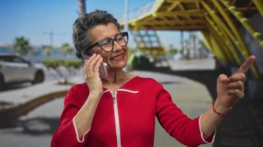 Gri saçlı, kırmızı elbiseli bir kadın telefonda konuşuyor. Şehir arka planında mutlu bir şekilde el kol hareketi yapıyor. Canlı şehir ortamını ve iletişimi öneriyor..