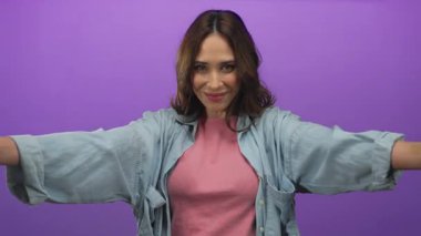 Kolları açık bir şekilde gülümseyen bir kadın, canlı mor bir zemin üzerinde, sıcaklığı ve pozitifliği iletiyor, günlük kıyafetleriyle esmer, yumuşak stüdyo ışıklarıyla aydınlatılıyor..