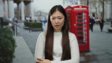 Şehirdeki uzun saçlı kadın kırmızı telefon kulübesinin yanında ciddi ifade ve el hareketi sergiliyor, şehir ortamında duygularını ve kültürel bağlamları aktarıyor..