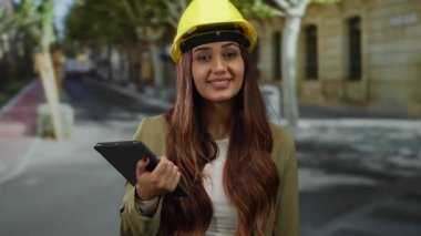 Kasket takan genç bir mimar güneşli bir sokakta açık hava ortamında profesyonel bir mühendisi resmeden bir tablet tutuyor..