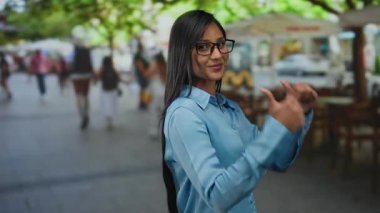 Mavi gömlekli genç bir kadın güneşli bir restoran sahnesinde canlı bir terasta bulanık insanlarla gülümserken başparmaklarını geriye doğru gösteriyor.