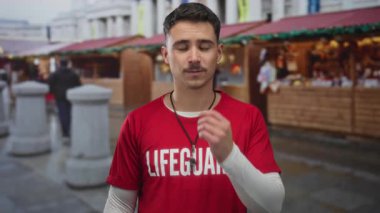 Kırmızı bir cankurtaran gömlekli genç İspanyol sokak arkaplanı ile bir açık markette duruyor, ahşap tezgahların ve taş sütunların yanında kapalı gözlerle bir öpücük savuruyor..