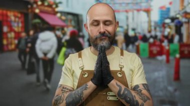 Bald man with tattooed arms and beard meditates with clasped hands in urban street setting, wearing brown apron and black gloves as people walk by in city environment.