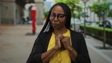 Kıvırcık saçlı ve gözlüklü bir kadın modern mimari ve yeşillikle şehir caddesinde el hareketi yapıyor..