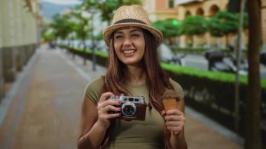 Güneşli bir günde şehir manzarasını keşfederken kamera şapkası ve dondurmayla gülümseyen kadın anılarını yakalıyor..