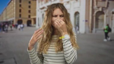 Bir şehir meydanında sarışın genç bir kalabalığın ortasında durmaktan tiksinerek el kol hareketi yapan bir kadın, bir şehir turizm bölgesinde tiksinti ifadesi sergiliyor..