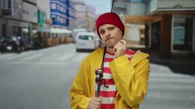 Çizgili tişörtlü ve sarı ceketli genç İspanyol bir adam elinde olta tutan bir şehir caddesinde duruyor, kırmızı bir şapka takıyor, açık havada düşünceli bir görünüm sergiliyor..