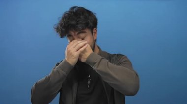 Sakallı, gülümseyen ve mavi arka planda yüzünü kapatan genç İspanyol adam neşe ve mutluluk gösteriyor..