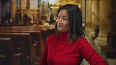 Kırmızı kazaklı, gülümseyen genç Çinli bir kadın dekoratif bir kilise ortamında el kol hareketi yapıyor. Düşünceli ve anlayışlı bir fikir..