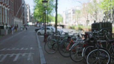 Amsterdam 'da bir kanal boyunca park etmiş bisikletler, arka planda ağaçlar, bir cadde ve binalar, insanların yürüdüğü sakin bir şehir manzarası yaratıyor..