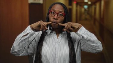 Gözlüklü bir kadın otel koridorunda neşeyle gülümsüyor, çizgili bir gömlek giyiyor, içeride neşe ve çeşitliliği somutlaştırıyor..