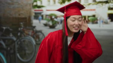 Kırmızı mezuniyet elbisesi giymiş bir kadın, kulaktan kulağa dinliyor, arka planda bisiklet ve binalarla bir şehir sokağında duruyor, neşe ve güveni ifade ediyor.